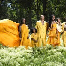 A family of six stands together in a sunlit field of soft white wildflowers, surrounded by lush green grass and tall, dark trees. At the center, a man and woman stand close, with four children gathered around them—one held in the woman’s arms, others standing confidently at their sides. All are dressed in coordinated, flowing and tailored outfits in vibrant shades of yellow and gold, creating a striking contrast against the deep green landscape. One figure on the left wears a dramatic, billowing gown that e
