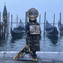 A masked figure in a Carnevale costume stands in front of gondolas and a dock at the edge of the island of Venice, Italy