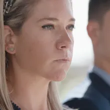 A young white woman with shoulder-length blond hair stares intently away from her; a young man in a suit is out of focus in the background