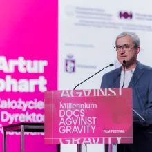 A middle-aged white man with grey hair and glasses wears a suit and a tee while standing at a podium that's branded as Millennium Docs Against Gravity in bright pink