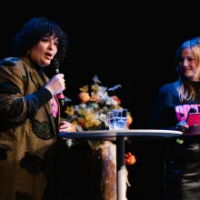 A light-skinned woman with shoulder-length curly black hair in a stylish printed coat holds a mic at a table with two glasses as a young blond-haired woman wearing a CPH:DOX tee stands in the background