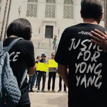 A peaceful outdoor rally taking place on the steps of Los Angeles City Hall. In the foreground, two people are seen from behind, wearing black t-shirts that read "Justice for Yong Yang." Behind them, a diverse crowd of participants of various ages, some wearing hats and sunglasses, hold signs that read "Mental Health Lives Matter" and "Koreans United for Justice."
