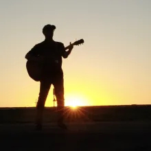 A lone man plays an acoustic guitar on the side of a freeway, silhouetted by the sunset at his back