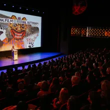 A young Black woman stands at a podium on a stage dwarfed by the audience in attendance with a sign for the International Rotterdam Film Festival behind her