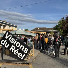 Under a crisp blue sky, a crowd of festivalgoers stands under the outdoor umbrellas. In front of them is a large sign that reads: "Visions du Reel."