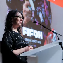 A middle-aged white woman with shoulder-length black wavy hair dons glasses and a black dress with white flowers at a podium with a mic