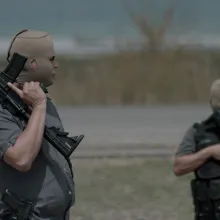 Two men in cammo gear stand in an empty field holding guns don skin-colored masks that reveal only their eyes, nose, and mouth