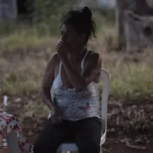 A dark skinned woman with dark curly hair in a messy bun wearing jeans and a ratty tank top sits on a plastic chair outside holds a lit cigarette up to her lips