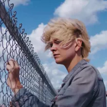  a young person with short, windblown blond hair standing beside a chain-link fence under a bright blue sky with large white clouds. They are wearing a gray denim-style jacket and appear to be looking through or past the fence with a quiet, reflective expression.