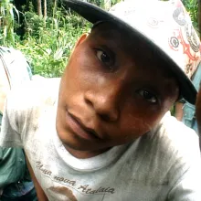 A man in a cap stares directly into the camera. The angle is distorted, as if shot with a fish eye lens. Another man in the background also looks into the camera. The two are in a densely vegetated area