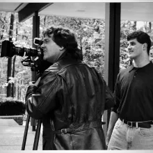 On the left, a man in a black leather jacket peers into a video camera. On the right, a younger man in a tucked polo observes.  Black and white photo