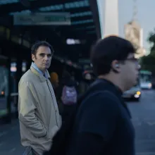 Middle-aged white man in a khaki rain coat over a light blue shirt stands in a somewhat crowded street looking straight at the camera