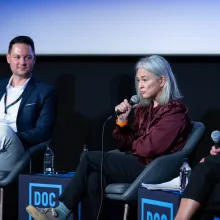 A woman in a maroon button-down shirt speaks into a microphone. She sits on a chair infront of a large screen. A man in a white shirt and blue blazer sits to her right. Both face forward. Tables on either side and in-between the two read "DOC NYC PRO"