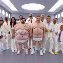 Two large men wear nothing but sneakers and diapers. One is decorated in tattoos, including a skull and crossbones and text which reads "BORN 2 DIE." Nine men and one woman stand behind the two in matching white suits. The ensemble appears in a sterile, grey room with futuristic, circular white lights