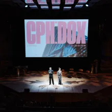 A man in a suit and a woman in a sequined gown stand in the center of a stage, illluminated by a spotlight. A large screen appears behind them which reads "CPH:DOX" in pink text