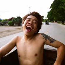 Young shirtless Asian man with dyed light red hair smiling broadly while on the bed of a moving truck on the road