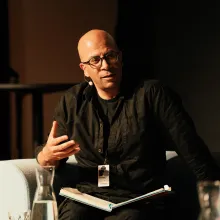 Bald man in glasses and dark brown collared shirt sits in white arm chair, gesturing with hand