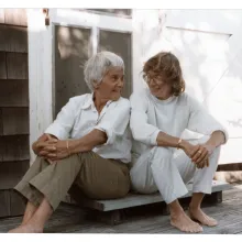 Two women sit on a doorstep smiling at each other. On the left is an older woman with short white hair in khaki pants and a white collared shirt. On the right is a woman with mid-length brown hair, glasses, a white turtleneck and white pants. Both are barefoot