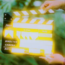 Two hands prepare to clap shut a slate in front of ferns. The text on the slate is obscured by shining yellow light