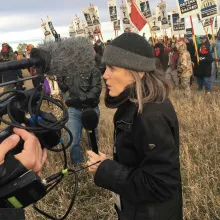 A middle-aged white woman in a black jacket and a grey wool hat stands in front of a protest holding a mic speaking into a camera