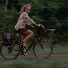 A woman with long grey hair, red glasses, a floral top, a short black skirt and sandals rides a black bike with a black bike bag along a forested path while looking to her right at the camera