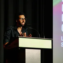 A man in a black shirt with black glasses and short dark hair and beard delivers a speech at a podium. Behind him is a black curtain and the lefthand side of a pink and green digital display showing text which cannot be read