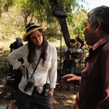 A woman (Lucrecia Martel) in a white button down shirt with glasses and a straw hat, wearing a walkie talkie and lanyard, speaks to a man in a red shirt. Behind the two are a film crew of three, one man holding a makeshift fill, one woman holding a boom pole and microphone and sound monitoring system, and one person mostly obscured by Martel