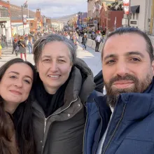 Selfie taken by a bearded man (right), woman with short grey hair (center), and woman with long, dark hair (left). All wear warm coats and smile. The background is a street lined with shops and pedestrians