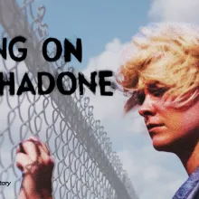 The image shows a young person with short, windblown blond hair standing beside a chain-link fence under a bright blue sky with large white clouds. They are wearing a gray denim-style jacket and appear to be looking through or past the fence with a quiet, reflective expression.  The composition feels cinematic and symbolic. The fence creates a strong visual barrier, suggesting themes of separation, confinement, longing, or being caught between two worlds. The open sky behind them adds contrast, giving the i