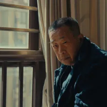 An aging Asian man with short-cropped hair in a bulky winter jacket glares at the camera while hunched over