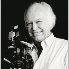 Black and white headshot of an older white balding man with tufts of white hair who smiles at the camera while holding a film camera