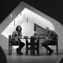 Two men sit at a table in a modern room framed by an unusually shaped doorway. Both look towards the camera. The man on the left has shoulder length dark hair and the man on the right has short greying hair and facial hair and glasses. Two mugs of coffee, a stack of books, and three decorative vases sit on the table. An abstract chandelier appears above the men's heads. Black and white image