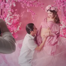 A photographer in frame right photographs a pregnant woman in a pink dress and pink flower crown in front of a pink background. A man in white kneels and kisses her stomach