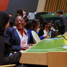 A candid photograph inside the United Nations General Assembly Hall captures two young delegates sharing a joyful moment between sessions. One student, wearing a black hijab, sits facing another delegate in a suit and conference lanyard, who is laughing openly. Around them, other students talk, move between seats, and prepare for proceedings, with microphones and the iconic General Assembly desks visible throughout the frame.  The image captures the heart of The Delegates: young people participating in seri