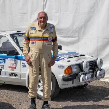 Brian Betteridge wearing a cream racing suit stands in front of his white Ford Escort RS1700T rally car with blue stripes and sponsor decals.