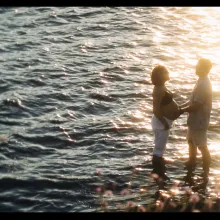 A beautiful, serene scene of a man and woman standing in the water at sunset as the man holds the woman's pregnant belly.