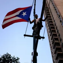 A man stands on a telephone pole with a large Puerto Rican flag flying next to him. Shot from beneath the pole, and the background is filled with the sky and one side of an apartment building