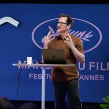 White middle-aged man with brown hair, glasses and a neat mustache wears a brown shirt against a blue backdrop at Cannes's Marche du Film standing in front of a laptop mid-performance
