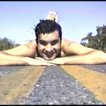 Grainy, digital photo of a smiling, shirtless person lying on the yellow painted lines of an asphalt road. Their chin rests on their hands and their feet appear to be raised behind their head