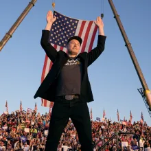 A man in a black suit jacket, grey T-shirt and black cap (Musk) stands smiling with arms raised above his head, looking to his left out of the corner of his eye. A massive American flag flies behind him from two cranes, hanging above a bleacher filled with people holding Trump Vance signs and a row of more American flags