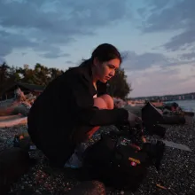A young woman squats on a beach holding a film camera at dusk