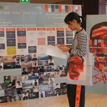 A woman in a striped shirt with a tote bag over her shoulder uses her smartphone. She stands in front of two large banners, one of which displays a schedule for the 2026 edition of What the Doc!, and one of which displays a graphic of a man's face (the same graphic which is on her bag) behind the words What the Doc! 2