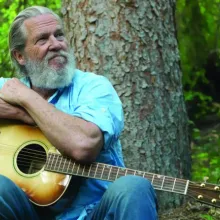 This image contains actor Jeff Bridges with a guitar, sitting in a forest looking up at the trees.
