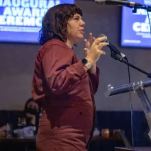 Jessica Devaney is a white woman with shoulder-length brown hair. She is wearing a rust jumpsuit and speaking into the mic at Doc 10's awards ceremony. Photo by Barry Brecheisen. 