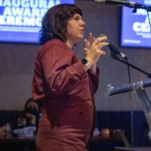 Jessica Devaney is a white woman with shoulder-length brown hair. She is wearing a rust jumpsuit and speaking into the mic at Doc 10's awards ceremony. Photo by Barry Brecheisen. 