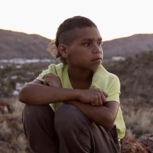 A young Dujuan Hoosan is sitting on a hill, looking pensively to his left. He is from the Arrernte Nation, group of Aboriginal Australian peoples from Central Australia.