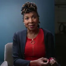 KIMBERLÉ CRENSHAW, Professor, UCLA and Columbia Schools of Law. She is an African-American woman, wearing a red blouse under a navy blue blazer; she is seated against a blue backdrop.