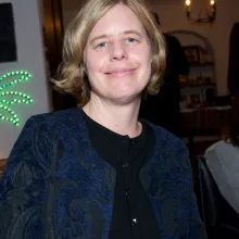 The late Beth Bird sits in a restaurant, smiling; she has short blond hair, and is wearing a blue jacket over a black shirt.