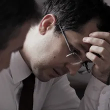 The image shows a close-up profile of a government official, sitting at a desk with his colleagues, examining documents. He has dark hair and glasses and is wearing a white shirt and a dark tie.