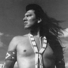 A Native American man, wearing traditional Indigenous accoutrements, stands in the middle of a plain, looking to his right. Courtesy of TBS